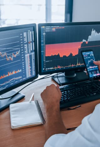 A person in front of three monitors examines stock trends
