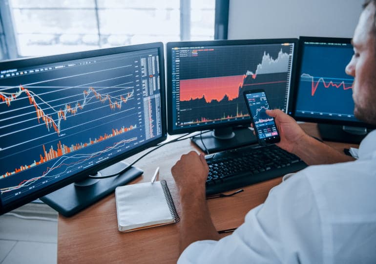 A person in front of three monitors examines stock trends